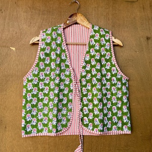 May include: A green quilted vest with a floral print and pink and white striped trim. The vest has a tie closure at the front.