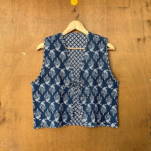 Puede incluir: Un chaleco azul con un estampado floral blanco. El chaleco está colgado en una percha de madera sobre un fondo de madera.