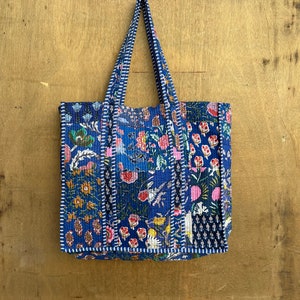 Puede incluir: Una bolsa de mano acolchada azul con un diseño de patchwork floral. La bolsa tiene dos asas y está colgada de una pared de madera.