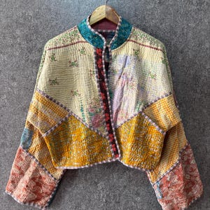 May include: A cropped jacket featuring a patchwork design with various floral and geometric patterns in shades of yellow, cream, teal, and orange. The garment has a mandarin collar, long sleeves, and a front opening with a dark floral trim.