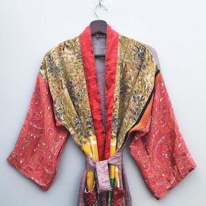 Könnte beinhalten: Ein farbenfroher, gemusterter Bademantel mit einem Paisley-Design in Rot-, Orange- und Gelbtönen. Der Bademantel hat einen roten Kragen und Besatz, mit einem Gürtel und Taschen. Der Bademantel hängt an einem Holzkleiderbügel.