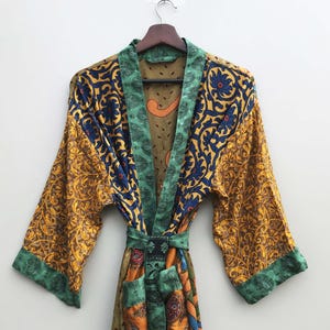 Może przedstawiać: Kolorowy, wzorzysty szlafrok z długimi rękawami wisi na drewnianym wieszaku. Szlafrok ma żółte, niebieskie, zielone i pomarańczowe tkaniny z kwiatowymi i paisley wzorami. Szlafrok jest wiązany w pasie pasującym paskiem.