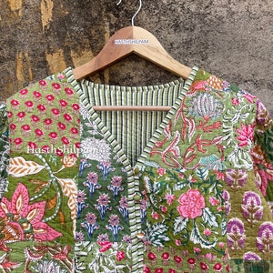 May include: A green quilted jacket with a floral print. The jacket has a V-neck and a button closure. The jacket is hanging on a wooden hanger with the text "HASTHSHILPAM" printed on it.