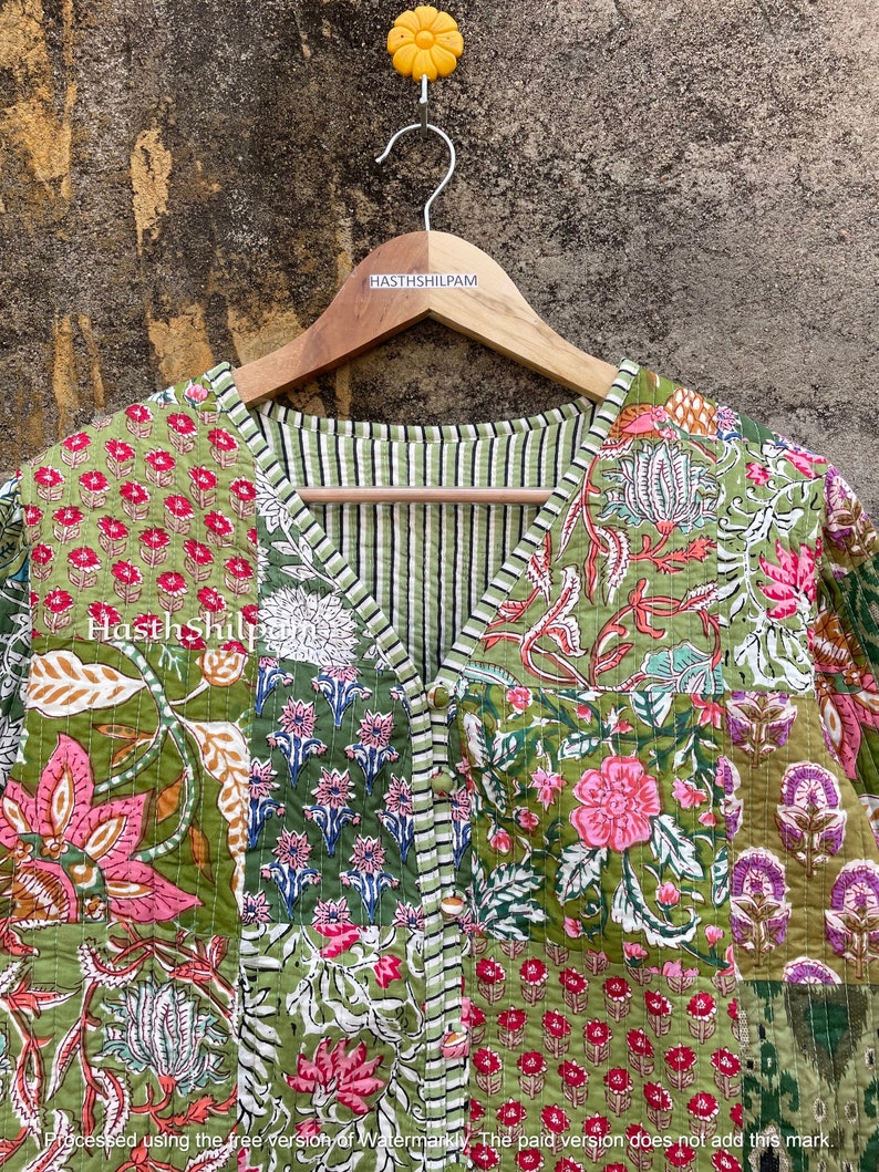 May include: A green quilted jacket with a floral print. The jacket has a V-neck and a button closure. The jacket is hanging on a wooden hanger with the text "HASTHSHILPAM" printed on it.