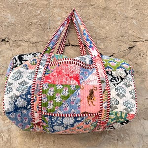 May include: A quilted duffel bag featuring a patchwork design of various floral and tiger prints in shades of red, green, blue, and pink. The bag has two top handles and is finished with a red and white striped fabric trim along the seams.
