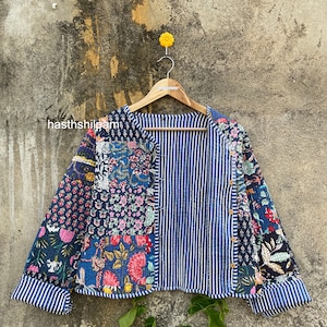 May include: A blue and white quilted jacket with a floral print. The jacket has a button closure and long sleeves. The jacket is hanging on a wooden hanger.