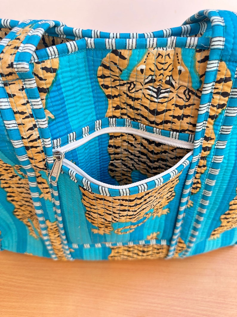 May include: A blue and white striped tote bag with a zippered pocket. The bag is made of quilted fabric and features a tiger print.
