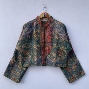 May include: A quilted jacket with a high collar and long sleeves. The jacket features a patchwork design with floral and geometric patterns in shades of green, blue, red, and gold. The jacket is hanging on a wooden hanger.