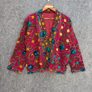 Könnte beinhalten: Eine leuchtend pinkfarbene, bestickte Jacke mit einem Blumenmuster in Gelb, Türkis und Grün. Die Jacke ist wendbar, mit einem rot-weiß gepunkteten Innenfutter. Sie hängt an einem Holzkleiderbügel.