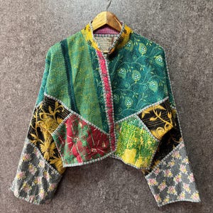 Peut inclure: Une veste courte confectionnée en tissu patchwork présentant divers motifs et couleurs, notamment du vert, du jaune, du rouge, du noir et du gris. Le vêtement possède un col officier, des manches longues et des surpiqûres visibles de style kantha.