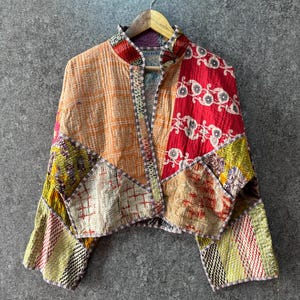 Op de afbeelding: Een kort jasje gemaakt van patchworkstof met een verscheidenheid aan kleuren en patronen, waaronder oranje, rood, geel en wit. Het kledingstuk heeft een hoge kraag, lange mouwen en zichtbare stiksels door het gehele doorgestikte materiaal.