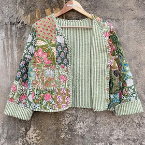 May include: A green quilted jacket with a floral print and striped lining. The jacket has a V-neck and is hanging on a wooden hanger.