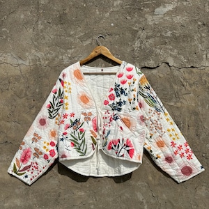 May include: A white quilted jacket with a floral print. The jacket has a V-neck and long sleeves. It is tied at the waist with a string.