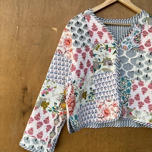 May include: A white quilted jacket with a patchwork design featuring floral and geometric patterns in shades of pink, blue, and green. The jacket has a V-neck and long sleeves with a striped trim.