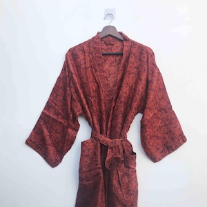 May include: A red floral patterned robe with wide sleeves hangs on a wooden hanger. The robe has a tie belt and pockets. The fabric has a rose pattern in a darker shade of red. The robe is displayed against a white background.