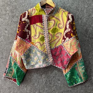 Könnte beinhalten: Eine kurz geschnittene Jacke mit einem Patchwork-Design aus verschiedenen Stoffen in Gelb, Rot, Grün und Grau mit feinen Kantha-Steppnähten. Das Kleidungsstück hat lange Ärmel, einen Stehkragen und eine Vorderöffnung mit einer Mischung aus floralen und abstrakten Mustern.
