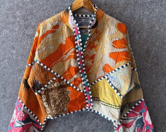 May include: A cropped jacket made from quilted fabric featuring a patchwork of orange, yellow, and pink floral and paisley patterns. The garment has long sleeves, a stand-up collar, and a black and white checkered trim along the edges and seams.