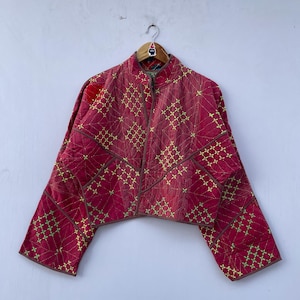 May include: A red quilted jacket with a stand-up collar and a zip closure. The jacket features a geometric pattern of yellow stitching and brown trim. The sleeves are wide and cropped, and the jacket is hanging on a wooden hanger.