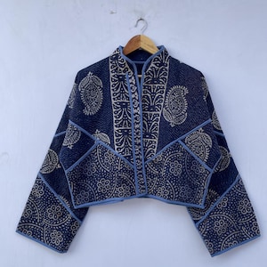 Puede incluir: Chaqueta azul marino con estampado de cachemir crema. La chaqueta corta tiene mangas largas y cuello mandarín, con ribetes azul claro. Se muestra en una percha de madera. El diseño de la chaqueta es un patchwork de tela estampada.