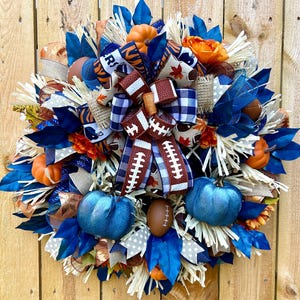 May include: A decorative wreath featuring a football theme. It includes blue and orange accents, miniature pumpkins, football-shaped ornaments, and decorative ribbons. The wreath is designed with a mix of textures and colors, perfect for fall or game day.