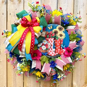 May include: A vibrant, circular wreath with a wooden background. It features a large red and yellow bow, colorful ribbons, and an assortment of artificial flowers. A central blue circle displays sunflowers, tulips, and red rain boots.