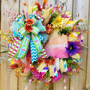 May include: A vibrant summer wreath with a wooden background. The wreath features colorful ribbons, faux flowers, and greenery. A sign in the center reads "Hello Summer" in white script. The wreath's color palette includes pink, yellow, orange, and turquoise.