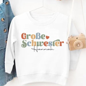 Puede incluir: Sudadera blanca de manga larga con el texto "Große Schwester Hannah" en letras coloridas. Un peto vaquero cuelga a la izquierda. Una pequeña cámara de madera cuelga a la derecha. Un logotipo de "Just 4 Every Child" está en la esquina inferior derecha.