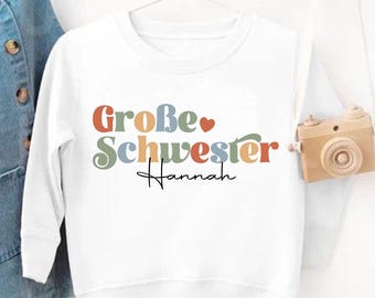 May include: White long-sleeved sweatshirt with the text "Große Schwester Hannah" in colorful letters. A denim overall hangs on the left. A small wooden camera hangs on the right. A logo for "Just 4 Every Child" is in the bottom right.