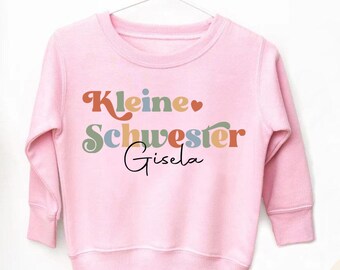May include: A light pink sweatshirt with the words "Kleine Schwester Gisela" in a retro font. The text is in various colors, including orange, green, and blue. The sweatshirt is hanging on a white hanger.