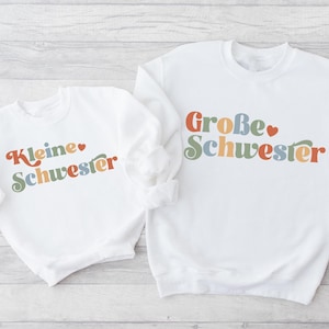 Puede incluir: Dos sudaderas blancas con texto de colores. La sudadera más grande dice "Große Schwester" con un corazón. La sudadera más pequeña dice "Kleine Schwester" con un corazón.