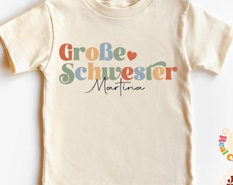May include: A light beige t-shirt with the text "Große Schwester Martina" in colorful, retro-style lettering. A small red heart is between the words "Große" and "Schwester".