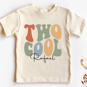 以下が含まれることがあります： クリーム色の半袖Tシャツ。レトロスタイルの文字で「TWO COOL」と書かれています。「Rafael」の文字が筆記体で下に書かれています。文字はオレンジ、緑、青、タンの色合いです。