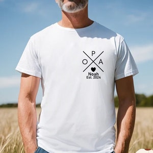 Peut inclure: T-shirt blanc avec un motif graphique noir en forme de croix avec les lettres "P", "O" et "A" à l'intérieur. Le motif comprend également un petit cœur et le texte "Noah Est. 2024".