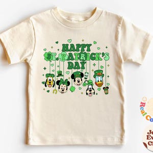 Puede incluir: Una camiseta beige claro con un diseño de trébol verde y el texto "Happy St. Patrick's Day". El diseño presenta a Mickey Mouse, Minnie Mouse, Donald Duck, Goofy y Pluto con sombreros verdes.