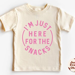 May include: A cream colored short-sleeved t-shirt with a pink circle graphic that says "I'm Just Here For The Snacks".