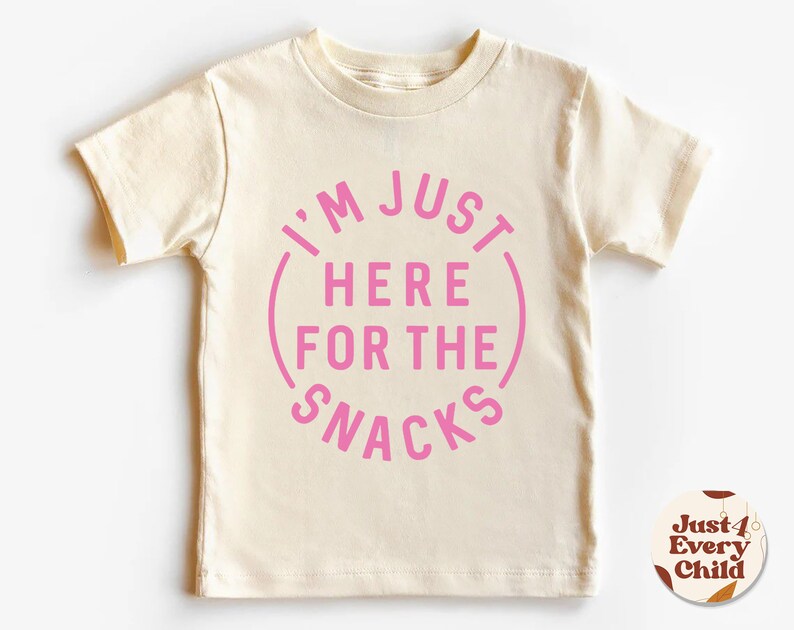 May include: A cream colored short-sleeved t-shirt with a pink circle graphic that says "I'm Just Here For The Snacks".