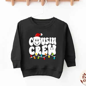 以下が含まれることがあります： 赤いサンタの帽子をかぶった笑顔の顔と「Cousin Crew」の文字が書かれた白いグラフィックが施された黒いクリスマスセーター。グラフィックはカラフルなクリスマスライトで囲まれています。