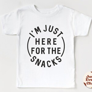 May include: White short-sleeved t-shirt with black text that reads "I'm Just Here For The Snacks" in a circle.