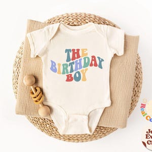 K&ouml;nnte beinhalten: Ein wei&szlig;er Baby-Strampler mit dem Text "The Birthday Boy" in Regenbogenfarben. Der Strampler liegt in einem geflochtenen Korb mit einer Holzrassel.