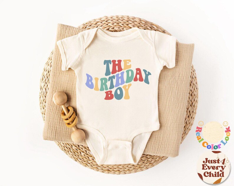 K&ouml;nnte beinhalten: Ein wei&szlig;er Baby-Strampler mit dem Text "The Birthday Boy" in Regenbogenfarben. Der Strampler liegt in einem geflochtenen Korb mit einer Holzrassel.