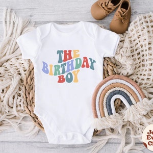 K&ouml;nnte beinhalten: Wei&szlig;er Baby-Strampler mit dem Text "THE BIRTHDAY BOY" in Regenbogenfarben. Der Strampler liegt auf einem geflochtenen Korb mit einem regenbogenfarbenen Seil daneben.