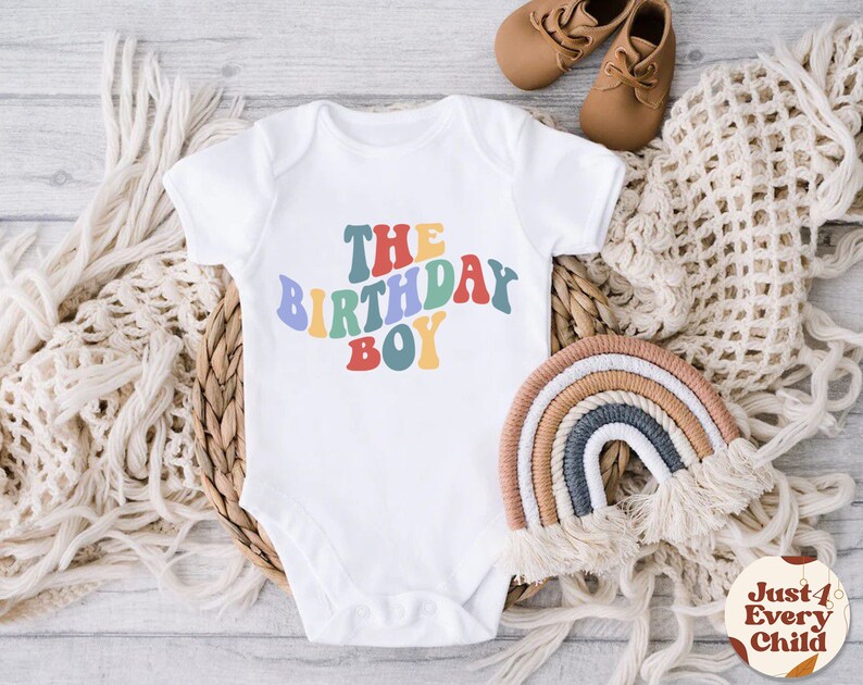 K&ouml;nnte beinhalten: Wei&szlig;er Baby-Strampler mit dem Text "THE BIRTHDAY BOY" in Regenbogenfarben. Der Strampler liegt auf einem geflochtenen Korb mit einem regenbogenfarbenen Seil daneben.
