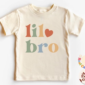 Peut inclure: Un t-shirt à manches courtes de couleur crème avec les mots "lil bro" en couleurs arc-en-ciel et un cœur rouge.