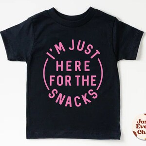 May include: Black short-sleeved t-shirt with pink text that reads "I'm Just Here For The Snacks".