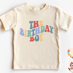 K&ouml;nnte beinhalten: Ein hellbeiges T-Shirt mit kurzen &Auml;rmeln und dem Text "The Birthday Boy" in Regenbogenfarben.