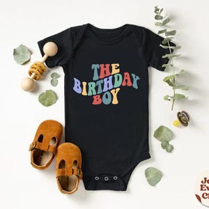 K&ouml;nnte beinhalten: Schwarzer Strampler mit dem Text "The Birthday Boy" in Regenbogenfarben. Der Text ist in einem Retro-Schriftart-Stil.
