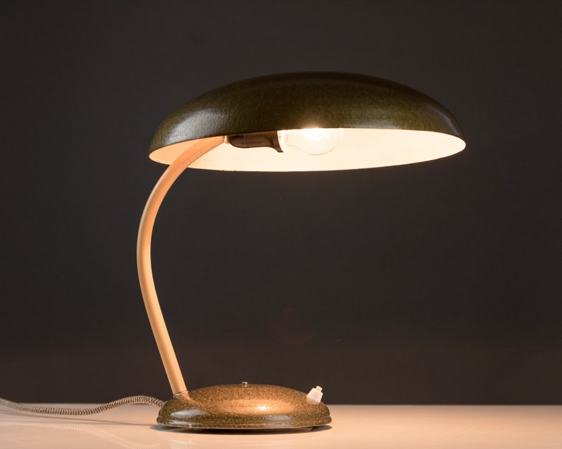 May include: A green metal desk lamp with a curved wooden arm and a white shade. The lamp is turned on and the light is shining.