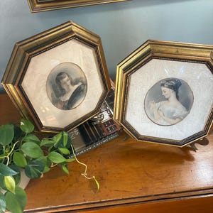 May include: Two antique gold framed portraits with oval images of women. The frames are octagonal with a gold finish. The portraits are in soft colors, with the women in period clothing. The frames are displayed on a wooden surface.