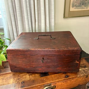 May include: A rectangular wooden box with a dark reddish-brown finish, featuring a metal handle attached to the top lid and a small keyhole centered on the front panel. The box has visible wood grain and signs of wear on its surface.