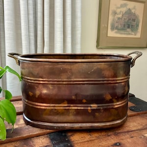 May include: An oval-shaped copper planter featuring a dark, weathered patina with visible horizontal ridges along the body. The vessel includes two curved metal handles attached to the sides, providing a functional grip for this metallic container.